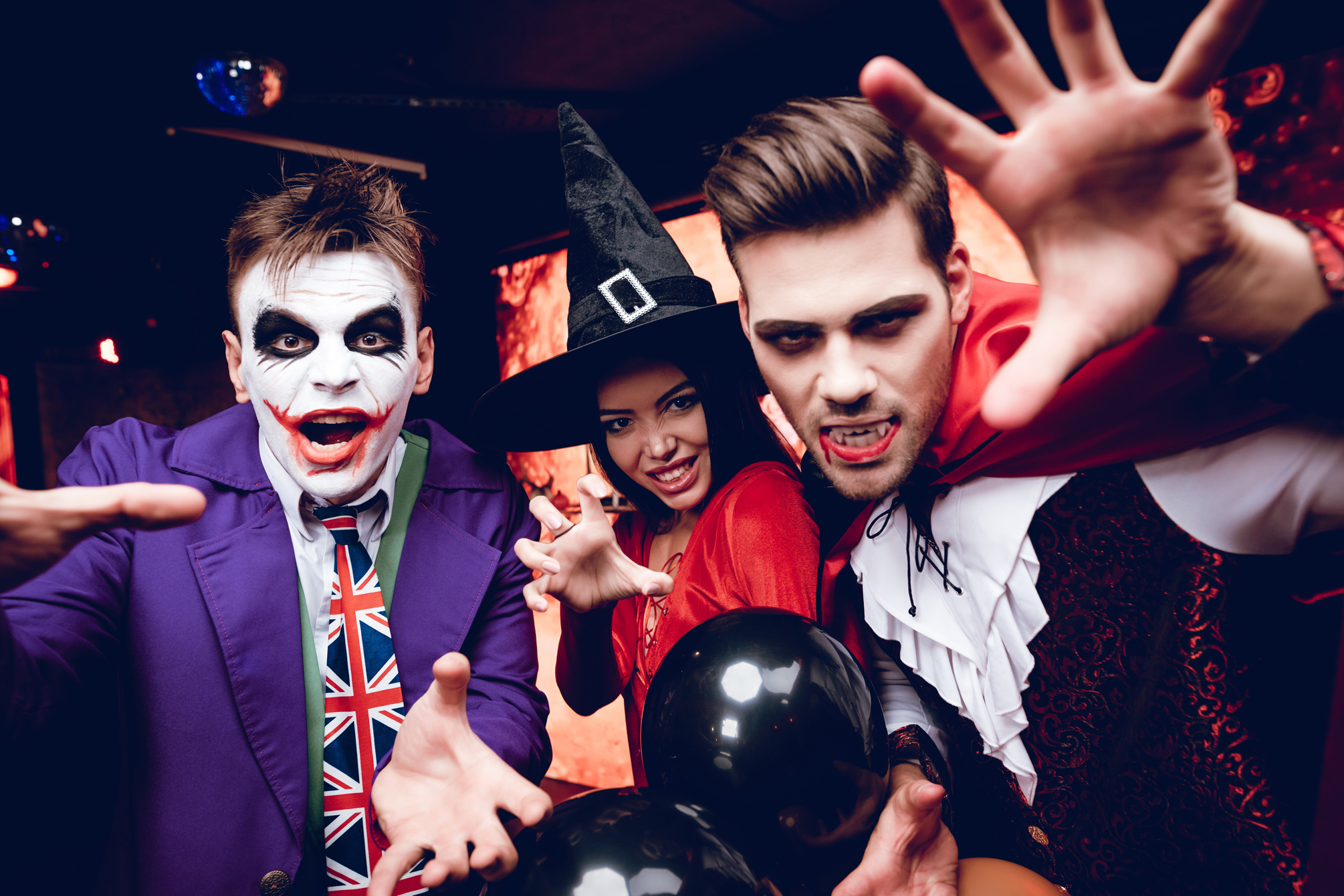 Three people in clown, witch and vampire costumes pose with hands reaching toward the camera.