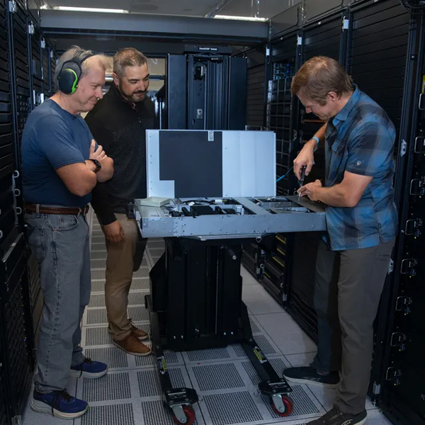 Three people inspect an open server chassis among equipment racks in a server room.